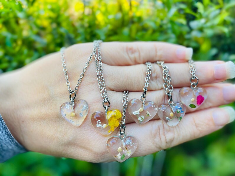 May include: Six silver necklaces with heart-shaped pendants filled with pressed flowers. The pendants are clear and show the flowers in detail. The necklaces are on a hand.