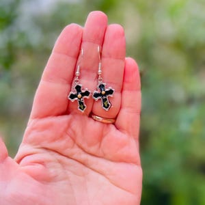 May include: A pair of silver cross earrings with black enamel and gold accents. The earrings are hanging from a person's hand.