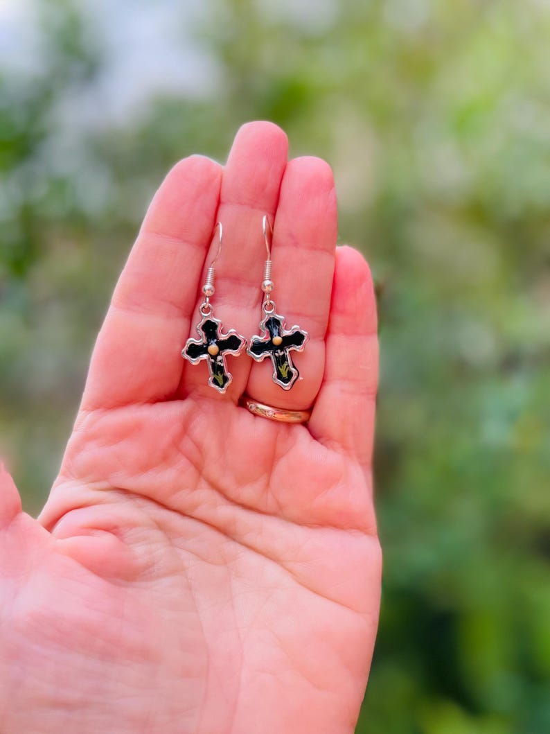 May include: A pair of silver cross earrings with black enamel and gold accents. The earrings are hanging from a person's hand.