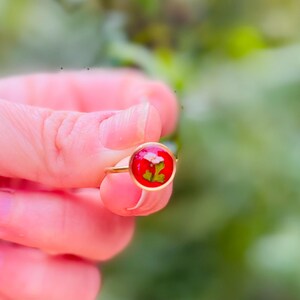 May include: A gold ring with a red resin setting featuring a pressed green leaf and white flower.