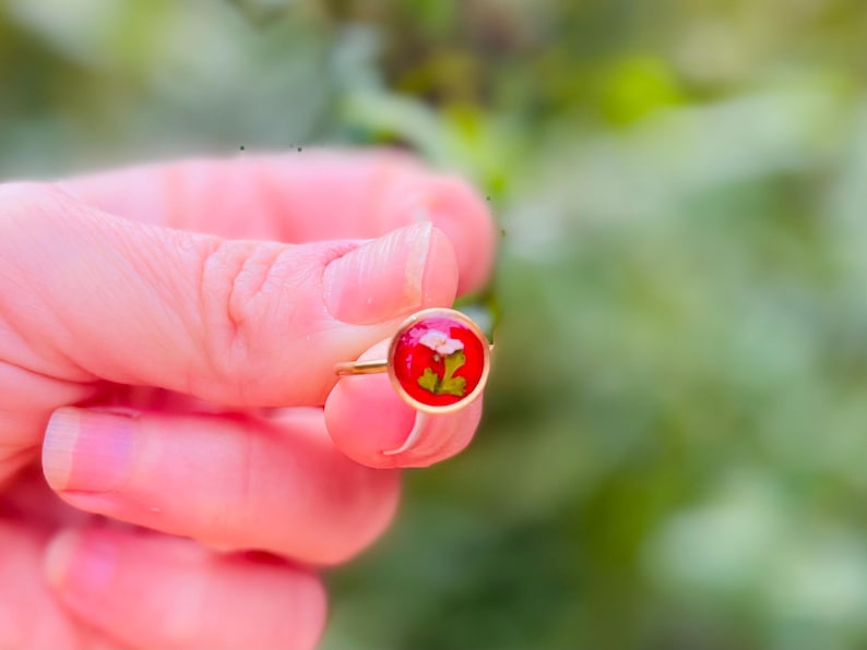 May include: A gold ring with a red resin setting featuring a pressed green leaf and white flower.