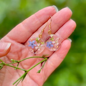 Puede incluir: Un par de pendientes de aro dorados con colgantes redondos de resina transparente. Cada colgante presenta flores azules y blancas prensadas.