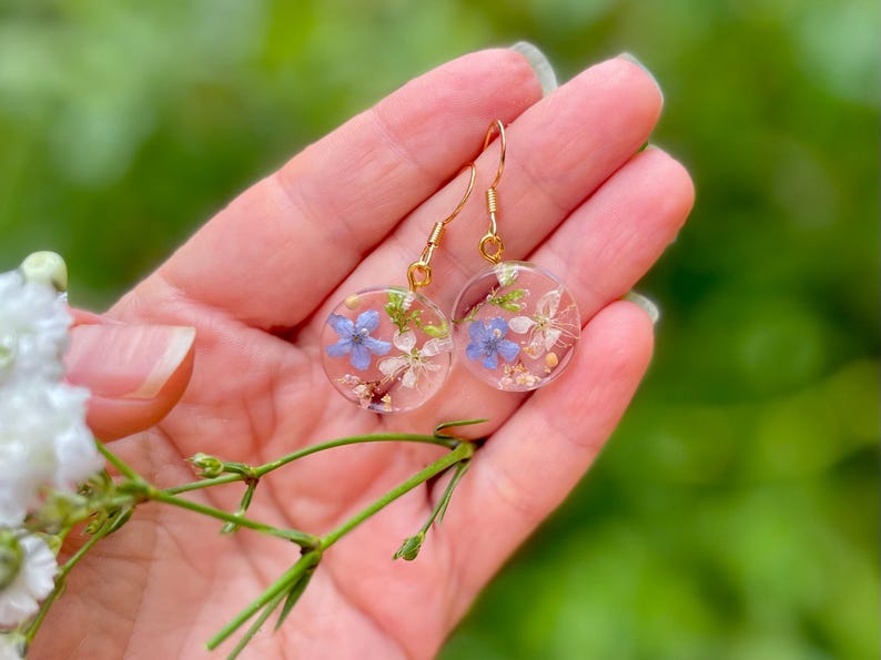Puede incluir: Un par de pendientes de aro dorados con colgantes redondos de resina transparente. Cada colgante presenta flores azules y blancas prensadas.