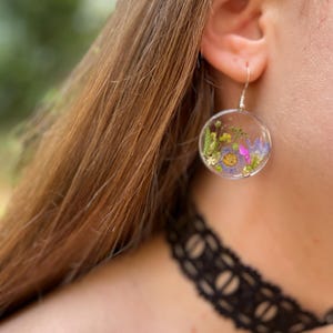 Puede incluir: Pendientes redondos y transparentes con flores secas encapsuladas en resina. Los pendientes presentan una variedad de flores coloridas, incluyendo moradas, amarillas y rosas, suspendidas en un entorno transparente. Cuelgan de ganchos plateados.