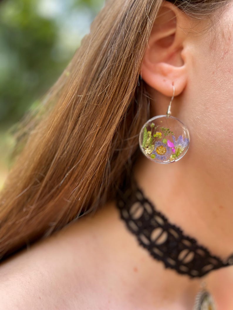 Puede incluir: Pendientes redondos y transparentes con flores secas encapsuladas en resina. Los pendientes presentan una variedad de flores coloridas, incluyendo moradas, amarillas y rosas, suspendidas en un entorno transparente. Cuelgan de ganchos plateados.