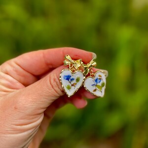 K&ouml;nnte beinhalten: Ein Paar goldene herzf&ouml;rmige Ohrringe mit wei&szlig;em Hintergrund und gepressten blauen und gr&uuml;nen Blumen. Die Ohrringe haben oben eine goldene Schleife.