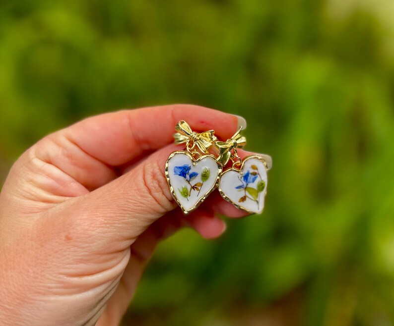 K&ouml;nnte beinhalten: Ein Paar goldene herzf&ouml;rmige Ohrringe mit wei&szlig;em Hintergrund und gepressten blauen und gr&uuml;nen Blumen. Die Ohrringe haben oben eine goldene Schleife.