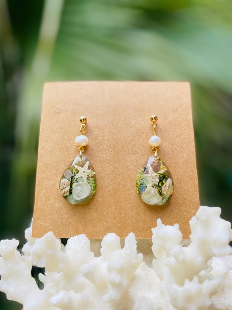 Op de afbeelding: Een paar druppelvormige oorbellen met goudkleurige stelen en kleine witte kralen. De doorzichtige hars oorbellen bevatten miniatuur schelpen, zeesterren en groen. De oorbellen worden gepresenteerd op een bruine kaart.