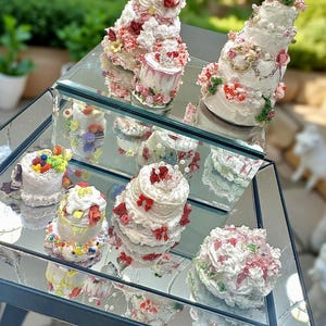 Puede incluir: Una colección de pasteles decorativos con glaseado blanco, adornados con detalles florales rojos, rosas y amarillos. Los pasteles se exhiben sobre una superficie espejada, reflejando los intrincados detalles y texturas de los postres.