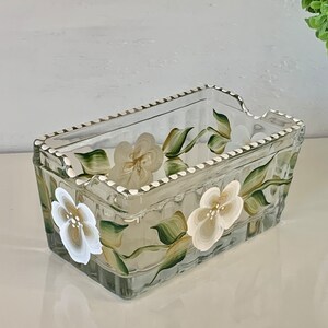 May include: A rectangular glass container with hand-painted white flowers and green leaves. The edges are trimmed with a white dotted pattern. The container is likely used for storage or display, adding a touch of floral elegance.