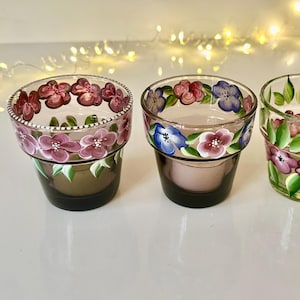 May include: Three hand-painted glass votive candle holders. Each is a different design, with floral patterns in shades of pink, purple, and blue, accented with green leaves. The candle holders are clear with a dark base.