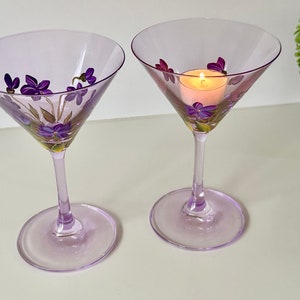 May include: Two lavender-colored martini glasses with hand-painted purple flowers. One glass has a small candle inside.