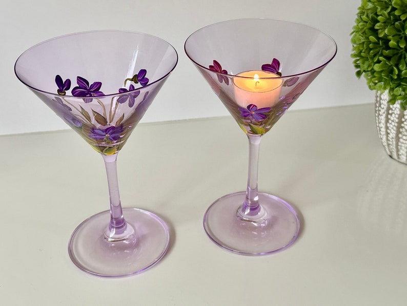 May include: Two lavender-colored martini glasses with hand-painted purple flowers. One glass has a small candle inside.