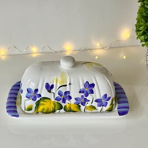 May include: White ceramic butter dish with a purple and white striped base. The lid features a floral design with yellow butterflies and purple flowers.
