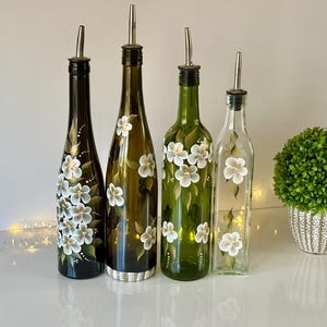 May include: Four glass bottles with hand-painted white flowers and green leaves. The bottles have silver pour spouts and are used for oil and vinegar.