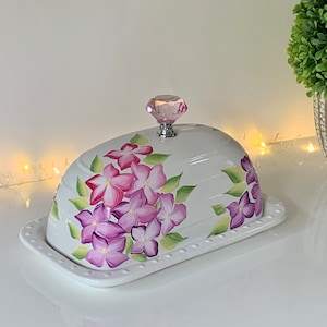 May include: White ceramic butter dish with a floral design featuring pink and purple flowers. The dish has a clear crystal knob on the lid.