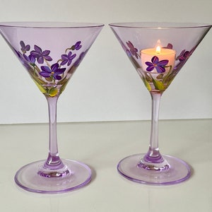 May include: Two lavender-colored martini glasses with hand-painted purple flowers and green leaves. One glass has a small white candle inside.