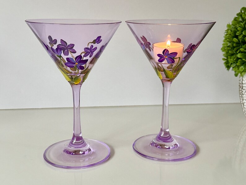 May include: Two lavender-colored martini glasses with hand-painted purple flowers and green leaves. One glass has a small white candle inside.