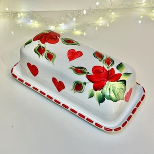 May include: White ceramic butter dish with a rectangular base and a domed lid. The dish is decorated with red roses, hearts, and green leaves. The rim is outlined with a red dotted pattern. A decorative kitchenware item.