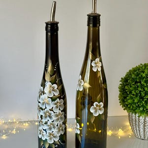 May include: Two glass bottles with white floral designs painted on them. The bottles have silver pour spouts and are filled with liquid.