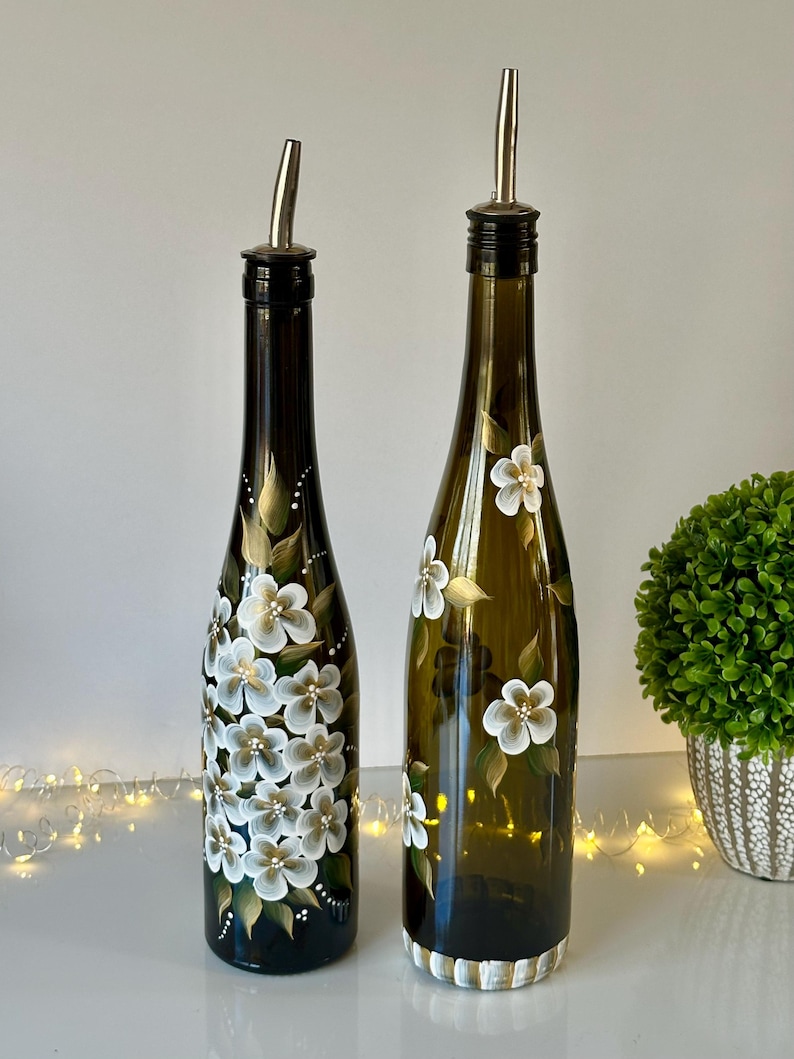 May include: Two glass bottles with white floral designs painted on them. The bottles have silver pour spouts and are filled with liquid.