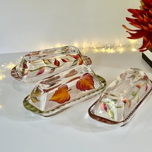 May include: Three glass butter dishes with painted autumn leaf designs. The dishes are clear glass with gold trim and feature painted leaves in shades of red, orange, and green.