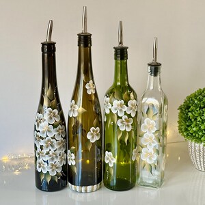 May include: Four glass bottles with hand-painted white flowers and green leaves. The bottles have silver pour spouts and are used for oil and vinegar.