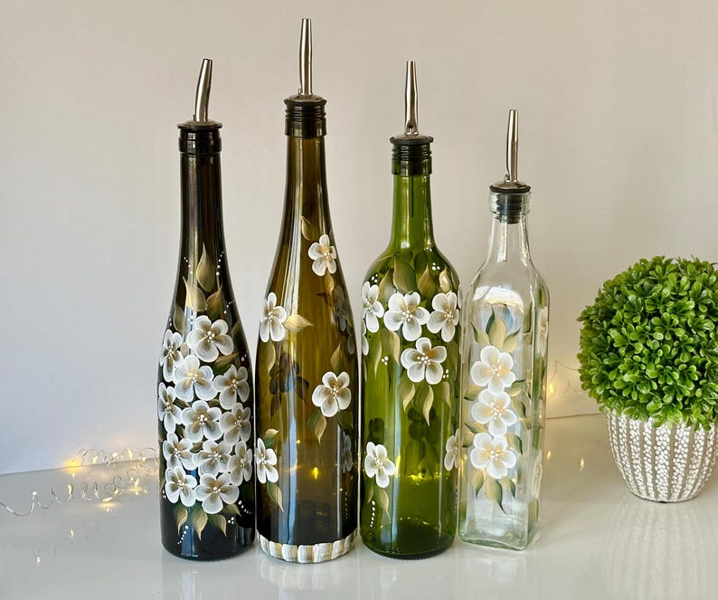 May include: Four glass bottles with hand-painted white flowers and green leaves. The bottles have silver pour spouts and are used for oil and vinegar.