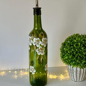 May include: A green glass bottle with a white floral design and a silver pour spout. The bottle is sitting on a white surface with a string of lights in the foreground.