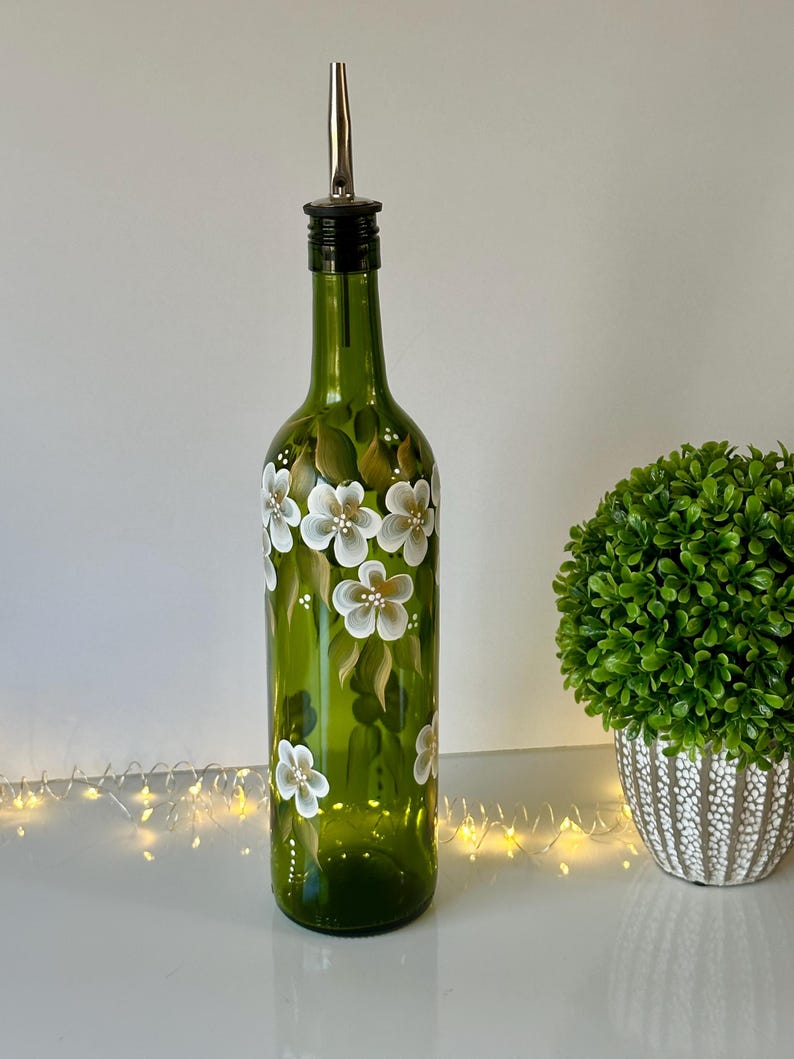 May include: A green glass bottle with a white floral design and a silver pour spout. The bottle is sitting on a white surface with a string of lights in the foreground.