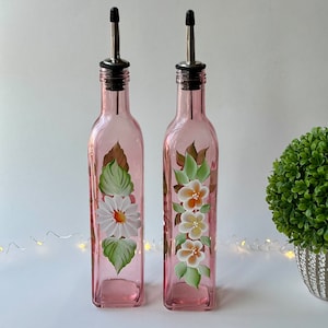 May include: Two pink glass bottles with hand-painted floral designs. The bottles have silver spouts and black tops. The bottles are on a white surface.