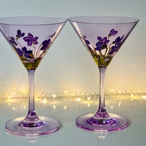 May include: Two purple glass martini glasses with hand-painted purple flowers. The glasses are on a white surface with a string of white lights in the background.