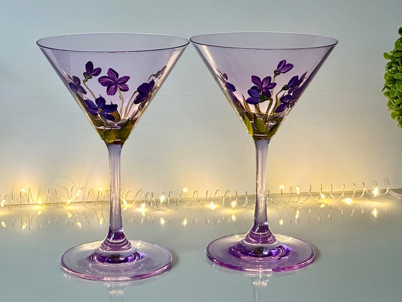May include: Two purple glass martini glasses with hand-painted purple flowers. The glasses are on a white surface with a string of white lights in the background.
