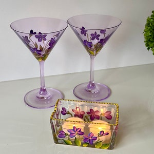 May include: Two purple glass martini glasses with hand-painted purple flowers and a clear glass votive holder with hand-painted purple flowers and two lit candles.