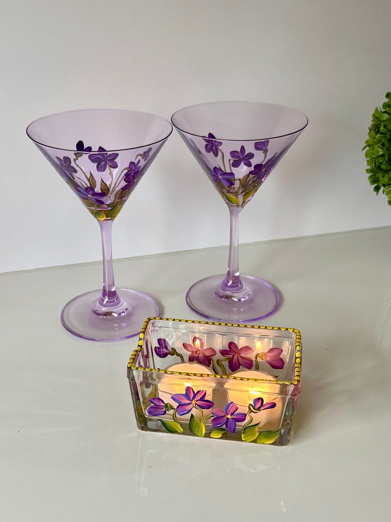 May include: Two purple glass martini glasses with hand-painted purple flowers and a clear glass votive holder with hand-painted purple flowers and two lit candles.