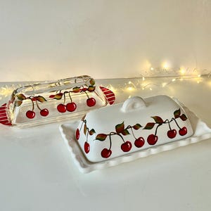 May include: Two butter dishes. One is clear glass with red and green cherry designs and red striped handles. The other is white ceramic with a matching cherry design and a lid. Both are rectangular.