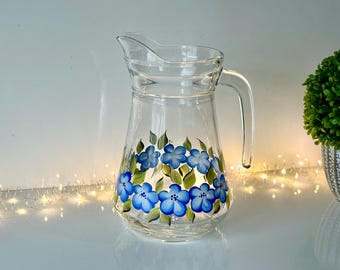 May include: A clear glass pitcher with a blue and green floral design. The pitcher has a wide base and a handle.