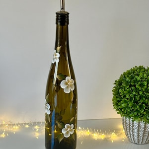 May include: A hand-painted olive oil bottle with a white floral design. The bottle is green glass and has a silver pour spout. The bottle is sitting on a white surface.