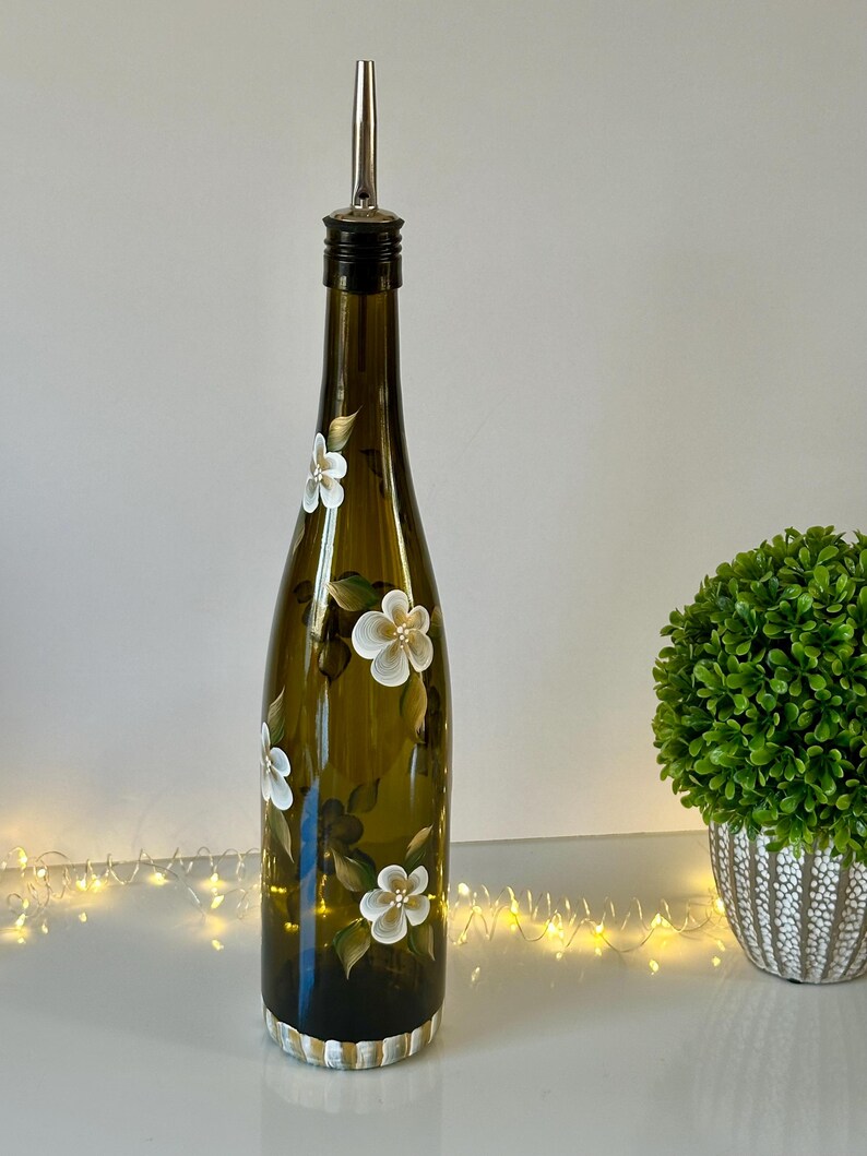 May include: A hand-painted olive oil bottle with a white floral design. The bottle is green glass and has a silver pour spout. The bottle is sitting on a white surface.