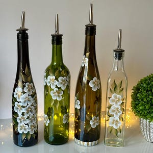 May include: Four glass bottles with white floral designs and silver pour spouts. The bottles are painted with white flowers and green leaves on a dark green, light green, brown, and clear glass background.