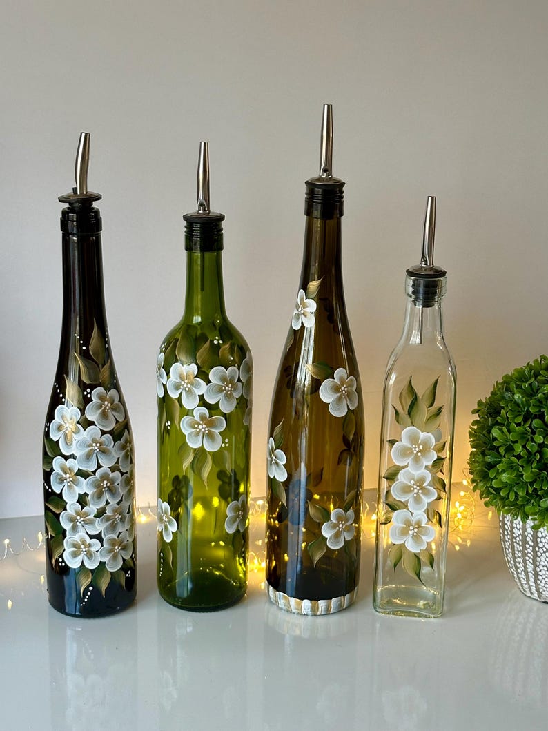 May include: Four glass bottles with white floral designs and silver pour spouts. The bottles are painted with white flowers and green leaves on a dark green, light green, brown, and clear glass background.