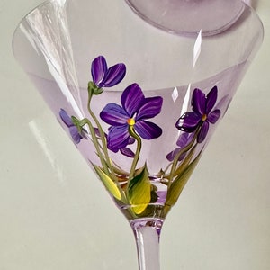 May include: A clear glass martini glass with a purple stem and a purple floral design. The flowers are painted on the inside of the glass and are purple with yellow centers.
