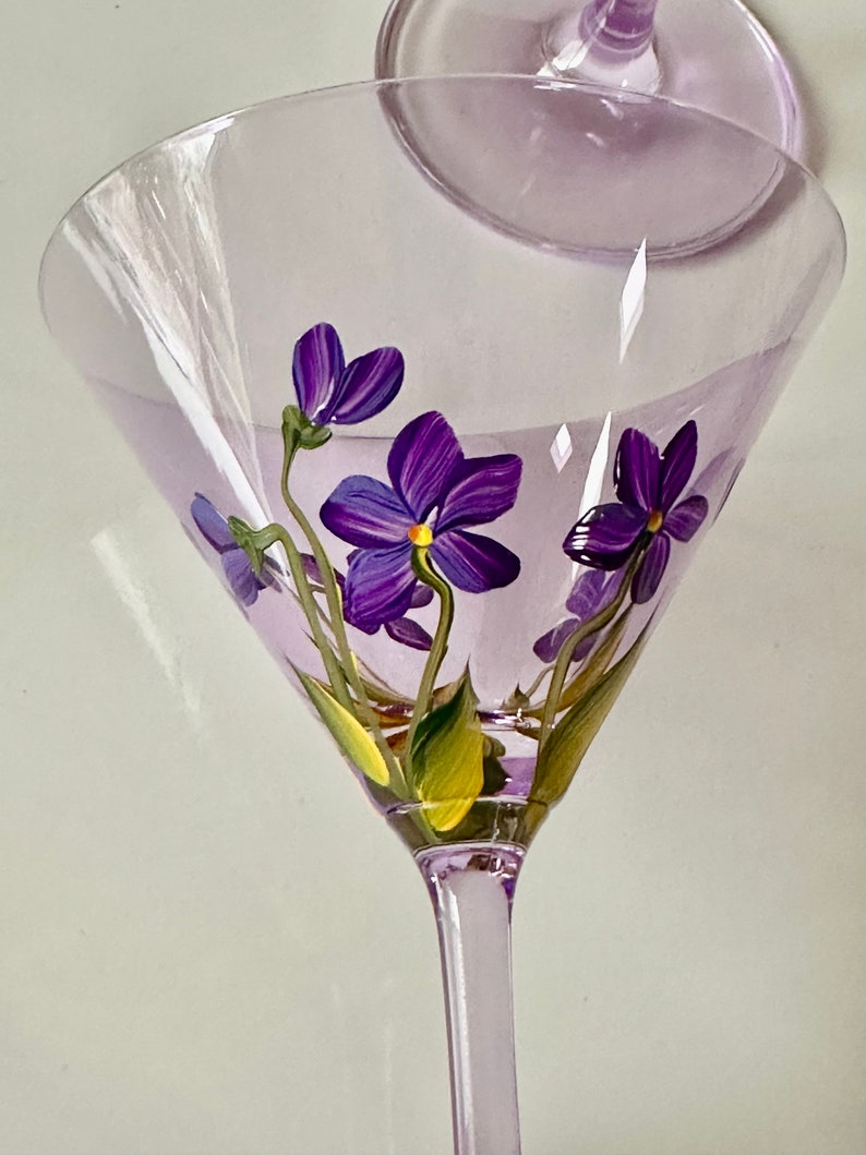 May include: A clear glass martini glass with a purple stem and a purple floral design. The flowers are painted on the inside of the glass and are purple with yellow centers.