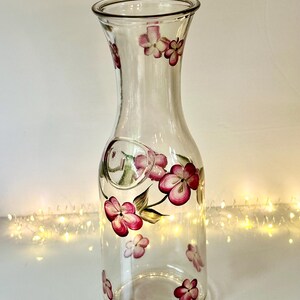 May include: A clear glass carafe featuring a hand-painted floral design with vibrant pink and magenta petals and green leaves. The vessel has a slender neck, a flared rim, and a rounded body, decorated with scattered blossoms around the exterior.