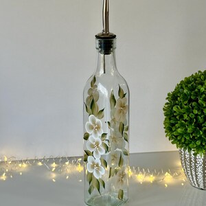 May include: A clear glass bottle with a silver pour spout and a hand-painted design of white flowers with green leaves. The bottle is sitting on a white surface with string lights.