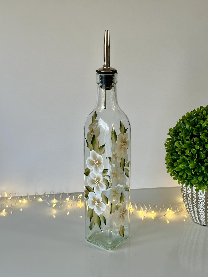 May include: A clear glass bottle with a silver pour spout and a hand-painted design of white flowers with green leaves. The bottle is sitting on a white surface with string lights.