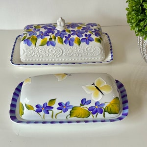 May include: Two ceramic butter dishes with floral designs. The top dish has a white base with purple flowers and a white knob. The bottom dish has a white base with purple flowers and a yellow butterfly.