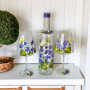 May include: A set of hand-painted wine glasses and a bottle with a floral design. The glasses and bottle feature purple and yellow flowers with green leaves.
