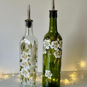 May include: Two glass bottles with white floral designs. The bottles have a clear glass body and a green glass neck. The bottles have a silver pour spout and a black stopper. The bottles are sitting on a white surface.