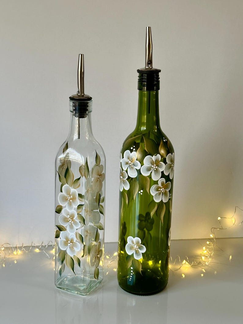 May include: Two glass bottles with white floral designs. The bottles have a clear glass body and a green glass neck. The bottles have a silver pour spout and a black stopper. The bottles are sitting on a white surface.